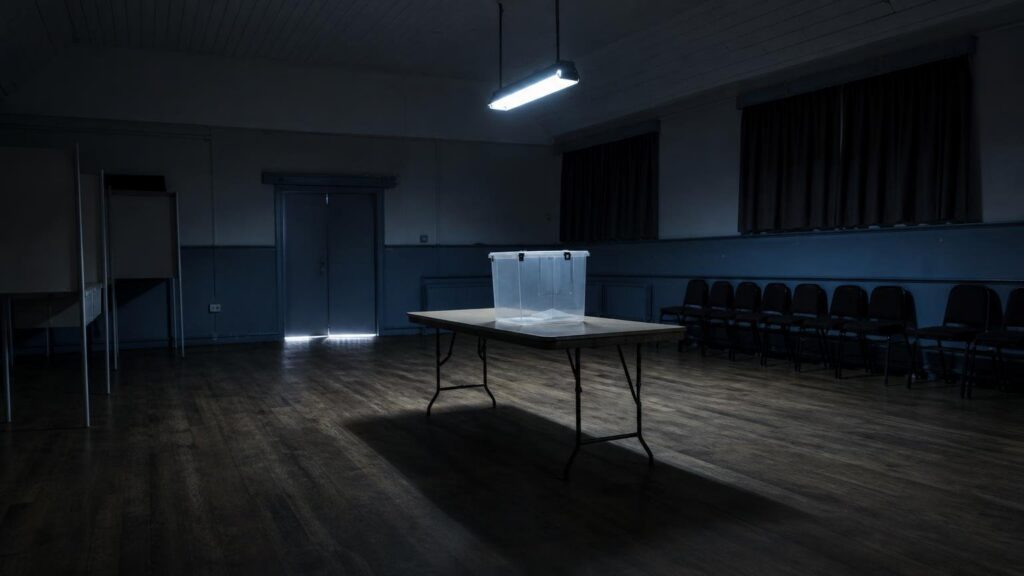 An empty polling station with a ballot box on a table and vacant observer chairs in dim light