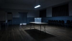 An empty polling station with a ballot box on a table and vacant observer chairs in dim light